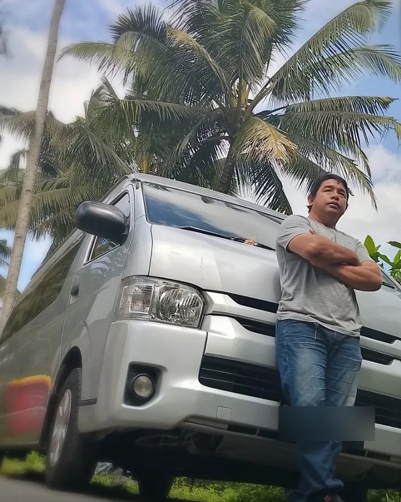 I Made Adi Suriawan standing beside his silver Toyota HiAce van under palm trees
