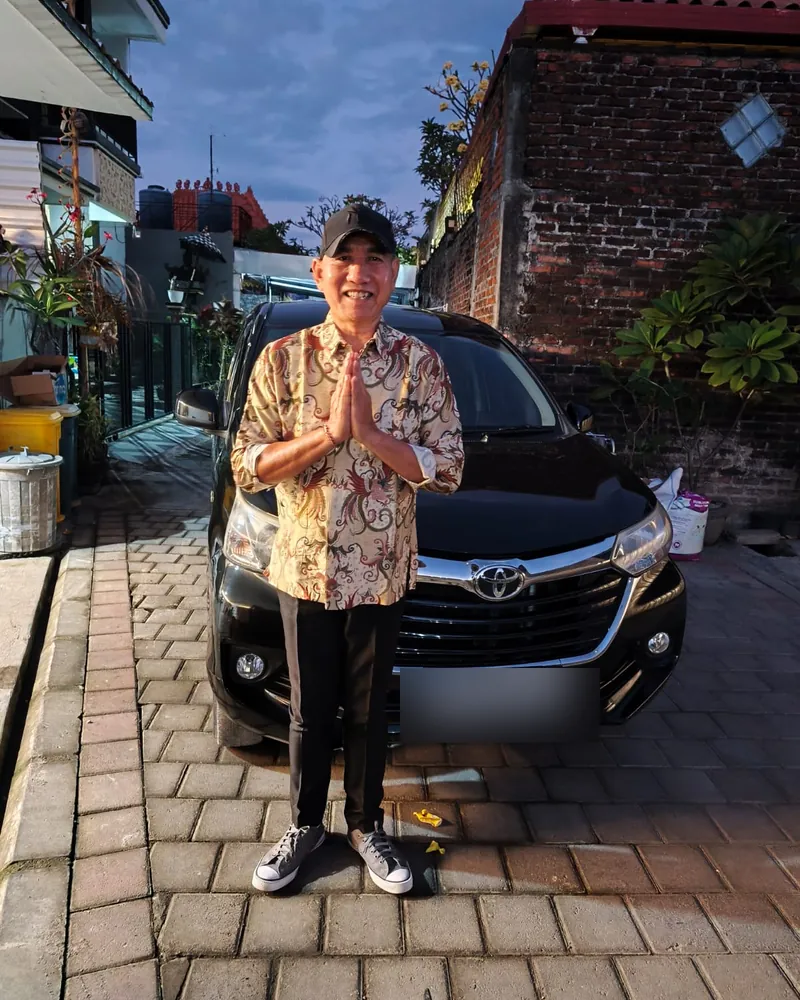 Ketut in a batik shirt, hands pressed together in greeting, beside his black Toyota Avanza
